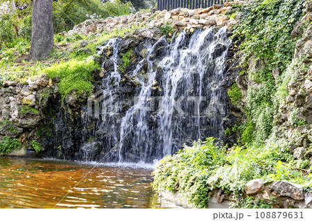 Waterfall. Waterfall in a park in Madrid. Artificial Mountain of Buen Retiro, called Mountain of the Cats. Dark stone background surrounded by green vegetation and plants and leaves. Autumn colors. Waterfall. Waterfall in a park in Madrid. Artificial Mountain of Buen Retiro, called Mountain of the Cats. Dark stone background surrounded by green vegetation and plants and leaves. Autumn colors. 108879631