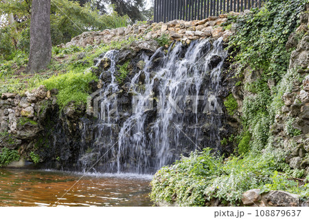 Waterfall. Waterfall in a park in Madrid. Artificial Mountain of Buen Retiro, called Mountain of the Cats. Dark stone background surrounded by green vegetation and plants and leaves. Autumn colors. 108879637