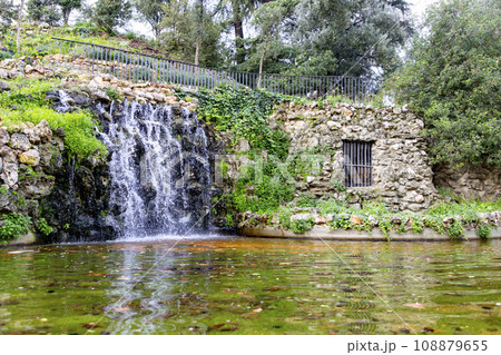 Waterfall. Waterfall in a park in Madrid. Artificial Mountain of Buen Retiro, called Mountain of the Cats. Dark stone background surrounded by green vegetation and plants and leaves. Autumn colors. 108879655