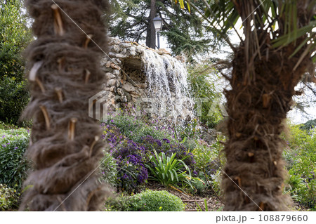 Waterfall. Waterfall in a park in Madrid. Artificial Mountain of Buen Retiro, called Mountain of the Cats. Dark stone background surrounded by green vegetation and plants and leaves. Autumn colors. Waterfall. Waterfall in a park in Madrid. Artificial Mountain of Buen Retiro, called Mountain of the Cats. Dark stone background surrounded by green vegetation and plants and leaves. Autumn colors. 108879660