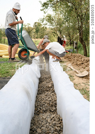 Man pours rubble from a wheelbarrow into a trench. Drainage works for drainage of ground water around the field. Man pours rubble from a wheelbarrow into a trench. Drainage works for drainage of ground water around the field. 108880035