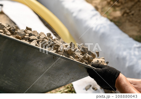 Drainage works for drainage of ground water. A man pours rubble crushed stone from a wheelbarrow into a trench close up. 108880043