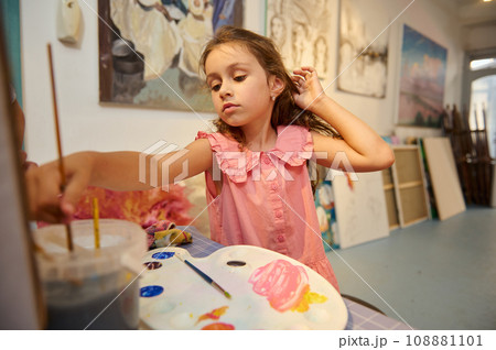 Caucasian little child girl dipping a paintbrush into water while painting with watercolors in creative art studio 108881101
