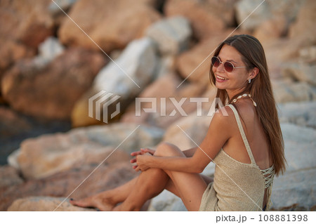 Closeup portrait of attractive brunette girl with long hair posing on rocky beach on sunset background. She wears elegant long dress, sunglasses. She is smiling. Closeup portrait of attractive brunette girl with long hair posing on rocky beach on sunset background. She wears elegant long dress, sunglasses. She is smiling. 108881398