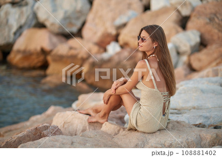 Closeup portrait of attractive brunette girl with long hair posing on rocky beach on sunset background. She wears elegant long dress, sunglasses. She is smiling. 108881402