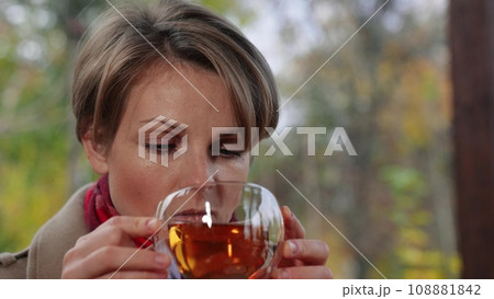 Cheerful lady drinks warm beverage while seated in gazebo with view of landscape. Lady takes break and enjoys hot drink. Lady with satisfied expression on face enjoys hot drink in peace with nature Cheerful lady drinks warm beverage while seated in gazebo with view of landscape. Lady takes break and enjoys hot drink. Lady with satisfied expression on face enjoys hot drink in peace with nature 108881842