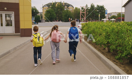 Junior children with schoolbags walk together after school lessons. Happy children with backpacks leave school yard joining hands. Little children go home after schooling in modern city street 108881857