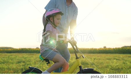 Diligent little girl in helmet rides bicycle with caring father in summer field Diligent little girl in helmet rides bicycle with caring father in summer field 108883311