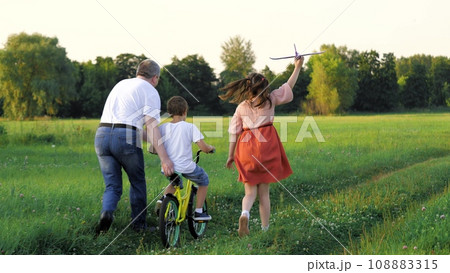 Father teaches little boy to ride bicycle while elder sister runs with plane Father teaches little boy to ride bicycle while elder sister runs with plane 108883315