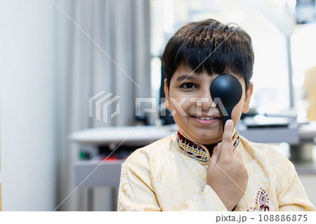 Happy Indian child boy examining eyesight modern ophthalmology equipment in clinic. 108886875