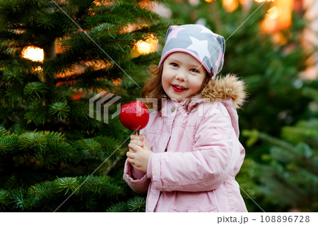 Cute little smiling preschool girl on German Christmas market. Happy child in winter clothes eating sweet sugared glazed xmas apple on with lights on background. Family, tradition, celebration concept 108896728
