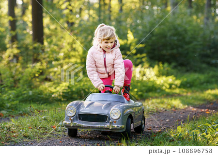 Little preschool girl driving big vintage toy car. Happy child having fun with playing outdoors. Active preschooler child enjoying warm autumn day in forest. Smiling stunning kid playing Little preschool girl driving big vintage toy car. Happy child having fun with playing outdoors. Active preschooler child enjoying warm autumn day in forest. Smiling stunning kid playing 108896758
