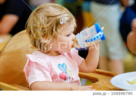 Adorable toddler girl drinking formula milk or water from bottle. Cute happy baby child taking food from dish at daycare or nursery canteen. 108896790