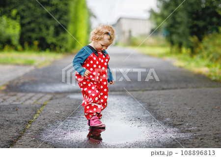 Little toddler girl wearing rain boots and trousers and walking during sleet, rain on cold day. Baby child having fun with jumping in a puddle. Active outdoors leisure and activity with little kids. Little toddler girl wearing rain boots and trousers and walking during sleet, rain on cold day. Baby child having fun with jumping in a puddle. Active outdoors leisure and activity with little kids. 108896813