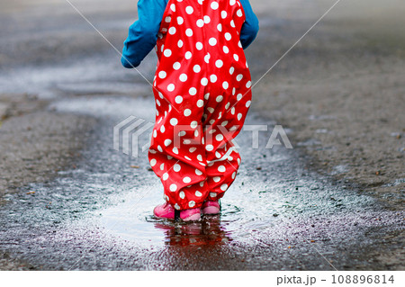 Little toddler girl wearing rain boots and trousers and walking during sleet, rain on cold day. Baby child having fun with jumping in a puddle. Active outdoors leisure and activity with little kids. Little toddler girl wearing rain boots and trousers and walking during sleet, rain on cold day. Baby child having fun with jumping in a puddle. Active outdoors leisure and activity with little kids. 108896814