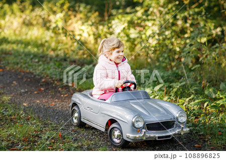 Little preschool girl driving big vintage toy car. Happy child having fun with playing outdoors. Active preschooler child enjoying warm autumn day in forest. Smiling stunning kid playing 108896825