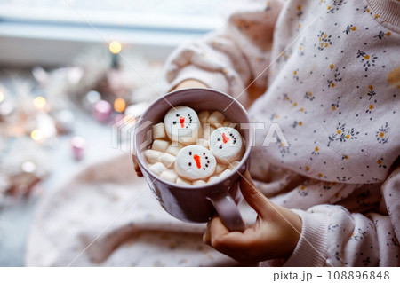 Unrecognizable little child girl holding cup with hot chocolate with marshmallows as snowman. Kid sitting near Christmas decorated window with lights. Unrecognizable little child girl holding cup with hot chocolate with marshmallows as snowman. Kid sitting near Christmas decorated window with lights. 108896848