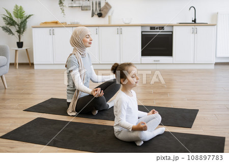 Peaceful woman in hijab meditating in seated asana with adorable girl indoors Peaceful woman in hijab meditating in seated asana with adorable girl indoors 108897783