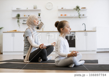 Peaceful woman in hijab meditating in seated asana with adorable girl indoors Peaceful woman in hijab meditating in seated asana with adorable girl indoors 108897784