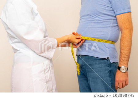 Nutritionist measuring overweight man's waist with tape in clinic, closeup Nutritionist measuring overweight man's waist with tape in clinic, closeup 108898745