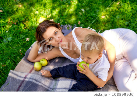 A young european mother with her cute little son lying on bed checkered plaid and smiling at camera, high angle . A child has a bitten green apple in his hand. The concept of parenting and spending A young european mother with her cute little son lying on bed checkered plaid and smiling at camera, high angle . A child has a bitten green apple in his hand. The concept of parenting and spending 108899936