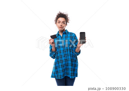 young smart curly brunette woman in blue plaid shirt shows plastic card and phone 108900330
