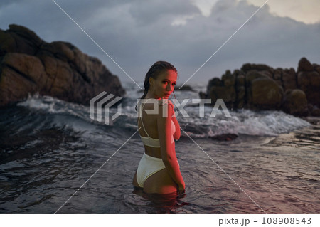 Attractive young woman enjoys nocturnal swim in ocean, illuminated by crimson light. Female beauty in stylish swimwear, night-time leisure at sea, tranquil waterscape surrounds. 108908543