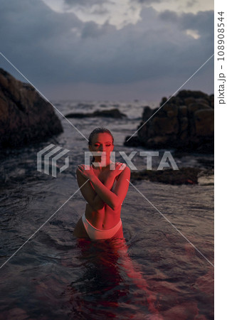 Attractive woman enjoys night swim in crimson lit ocean. Fashionable female basking in red illumination, sea backdrop, tranquil evening setting. Attractive woman enjoys night swim in crimson lit ocean. Fashionable female basking in red illumination, sea backdrop, tranquil evening setting. 108908544