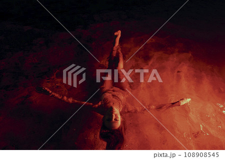 Ethereal woman floats tranquilly in lagoon illuminated by crimson light, nighttime serenity enveloping her form. Feminine grace drifts gently, waterscape aglow in ruby tones providing surreal setting. 108908545