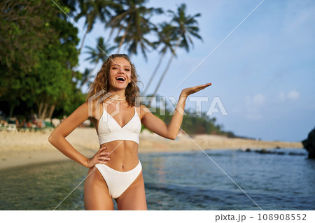 Cheerful woman in bikini presenting with hand on beach. Smiling female model showcases product placement outdoor. Summer beachwear fashion, tropical coastline lifestyle. Advertising, marketing concept 108908552
