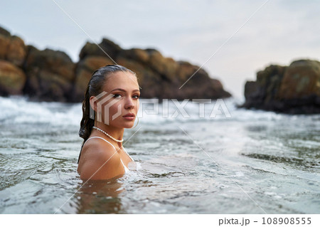 Woman with waterproof makeup submerges in ocean, stones backdrop. Female tours seascape, enduring swimwear stylish jewelry. Adventurous traveler explores coastal region, water resistant cosmetics. Woman with waterproof makeup submerges in ocean, stones backdrop. Female tours seascape, enduring swimwear stylish jewelry. Adventurous traveler explores coastal region, water resistant cosmetics. 108908555