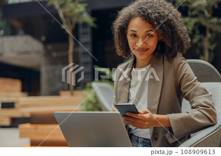 Positive female freelancer holding mobile phone while sitting in coworking and work on laptop Positive female freelancer holding mobile phone while sitting in coworking and work on laptop 108909613