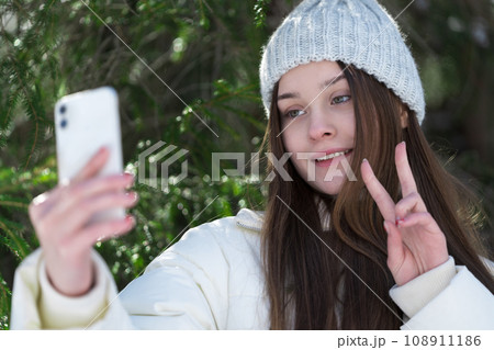 Portrait cheerful teenager girl using smartphone, showing gesturing peace sign, sending video selfie 108911186