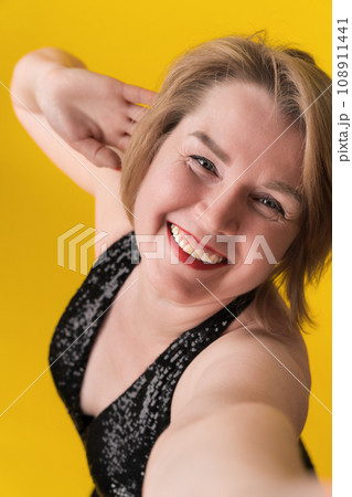Portrait of happy adult blonde in party dress, stretching hand to take selfie, looking at camera 108911441