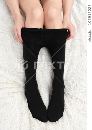 Closeup view of woman hands dressing black tights over legs carefully. Crop view woman legs on bed 108913028
