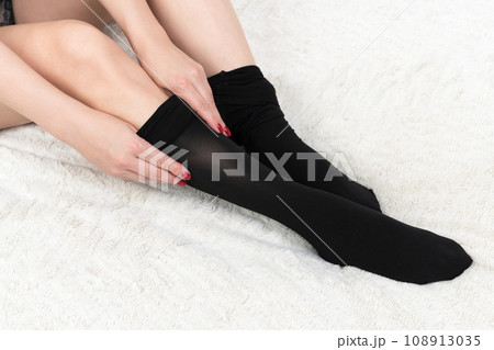 Close-up of woman hands putting on black pantyhose over legs carefully. Crop view of female on bed 108913035