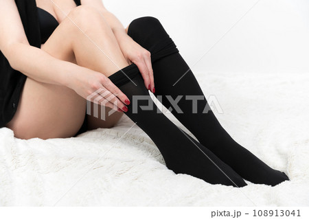 Woman putting on black pantyhose over legs. Cropped view of unrecognizable female sitting on bed 108913041