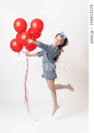 Playful girl in striped dress is jumping, holding bunch of red balloons in hand, looking at camera Playful girl in striped dress is jumping, holding bunch of red balloons in hand, looking at camera 108913279