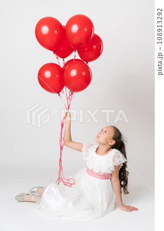 Smiling girl dressed in long white dress holding lot party red balloons in hand, looking up at balls Smiling girl dressed in long white dress holding lot party red balloons in hand, looking up at balls 108913292