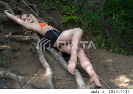 Slim woman with long legs lying down on ground on tree roots in forest, dressed in bikini top, skirt Slim woman with long legs lying down on ground on tree roots in forest, dressed in bikini top, skirt 108914352