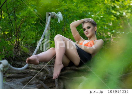 Sensual adult woman lying down and poses on ladder made of tree roots in summer forest 108914355