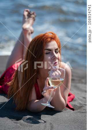 Woman lies on sandy beach in red evening dress with glass of white wine in hand. High angle shot 108914741