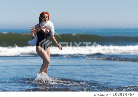 Smiling redhead woman in black swimsuit stands ankle deep in waves of ocean, holding glass of wine Smiling redhead woman in black swimsuit stands ankle deep in waves of ocean, holding glass of wine 108914767