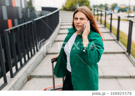 Young unhappy redhead businesswoman with red suitcase making phone call over mobile phone in the city. Portrait of a furious woman yelling at smartphone. Negative people emotion. 108925578