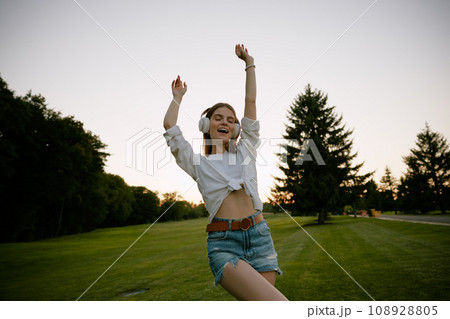 Happy teenage woman wearing headphones dancing on park lawn 108928805