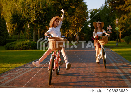 Happy funny young girl with female friend riding on bicycle with raised legs Happy funny young girl with female friend riding on bicycle with raised legs 108928889