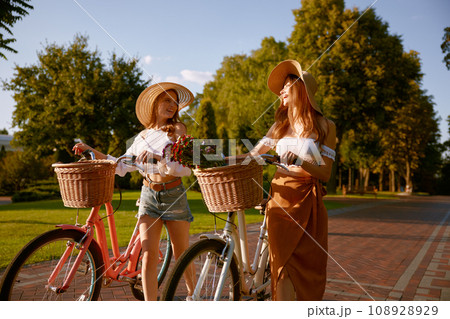 Two girl best friends nice conversation while walking with bicycle at park Two girl best friends nice conversation while walking with bicycle at park 108928929