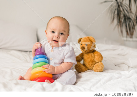 Cute baby playing with colorful rainbow toy pyramid and teddy bear sitting on bed in white sunny bedroom copy space. Toys for little kids. Child with educational toy. Cute baby playing with colorful rainbow toy pyramid and teddy bear sitting on bed in white sunny bedroom copy space. Toys for little kids. Child with educational toy. 108929897