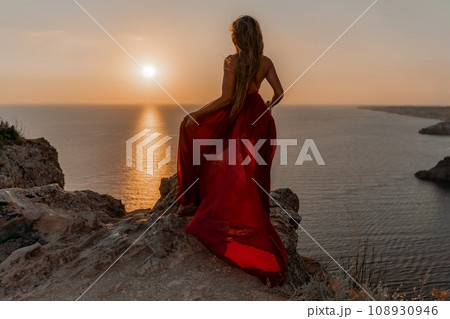 Woman sunset sea red dress, side view a happy beautiful sensual woman in a red long dress posing on a rock high above the sea on sunset. Woman sunset sea red dress, side view a happy beautiful sensual woman in a red long dress posing on a rock high above the sea on sunset. 108930946