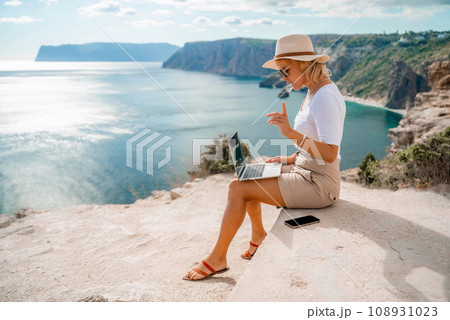 Freelance women sea working on the computer. Good looking middle aged woman typing on a laptop keyboard outdoors with a beautiful sea view. The concept of remote work. 108931023